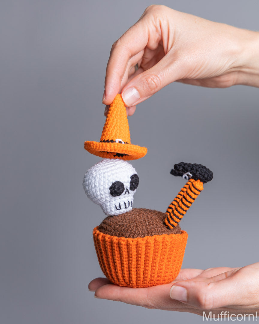A handcrafted crocheted Halloween cupcake decoration held in two hands against a solid gray background. The decoration features an orange cupcake liner, brown frosting, a white skull, an orange witch hat, and a striped orange and black candle.