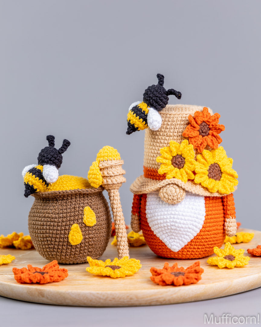 Crocheted bee and gnome figurines styled with sunflowers, honey pots, and a wooden honey dipper on a light wood tray against a solid gray background.