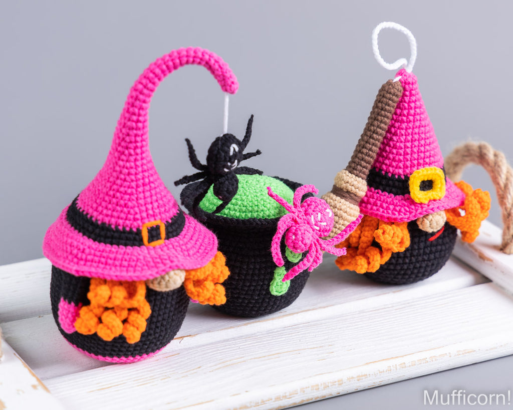 Three crocheted Halloween ornaments shaped like witch hats over black cauldrons, displayed on a white wooden surface against a gray background. One ornament has a pink hat with a gold buckle, one has a green cauldron with a black spider, and one has a pink hat with a broom handle.