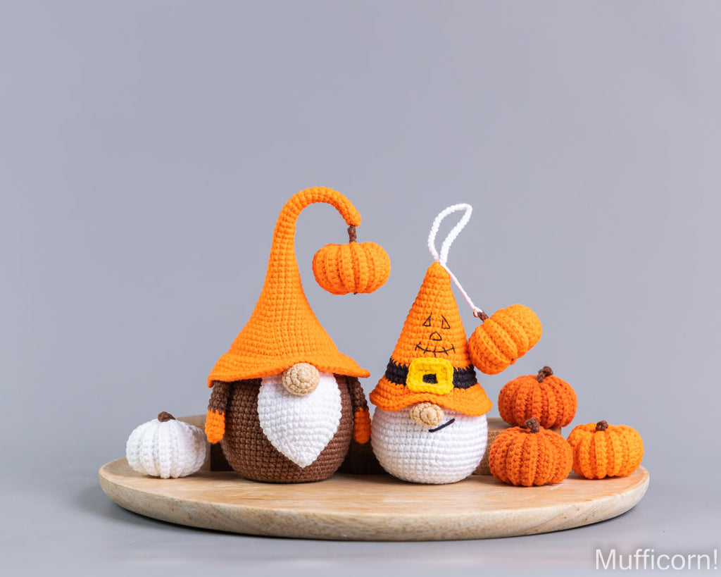 Two crocheted Halloween gnomes on a round wooden tray, surrounded by small orange and white pumpkins, against a solid gray background.