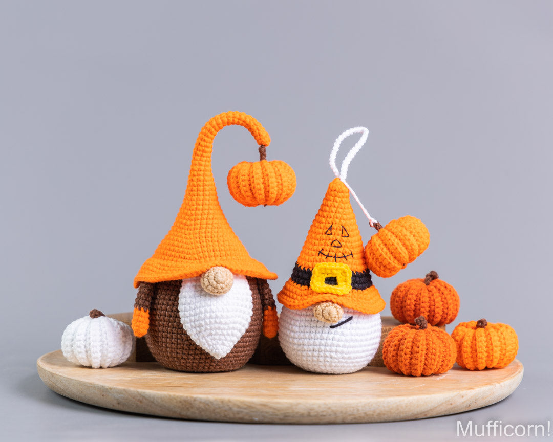 Two crocheted Halloween gnomes with orange hats and white/cream faces, displayed on a round wooden tray with small orange and white pumpkins against a plain gray background.