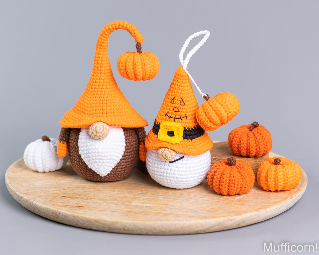 Two crocheted autumn gnomes with orange and white hats, brown and white bodies, and small orange pumpkins, displayed on a round wooden board against a gray background.