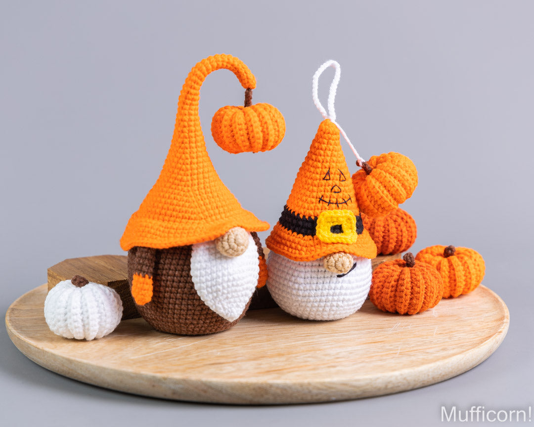 Two crocheted autumn gnome ornaments on a round wooden tray, surrounded by small crocheted pumpkins, against a solid gray background.