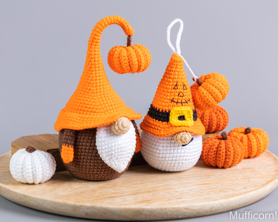 Two crocheted Halloween gnomes with orange pointed hats and small pumpkin decorations, displayed on a round wooden tray against a neutral gray background.