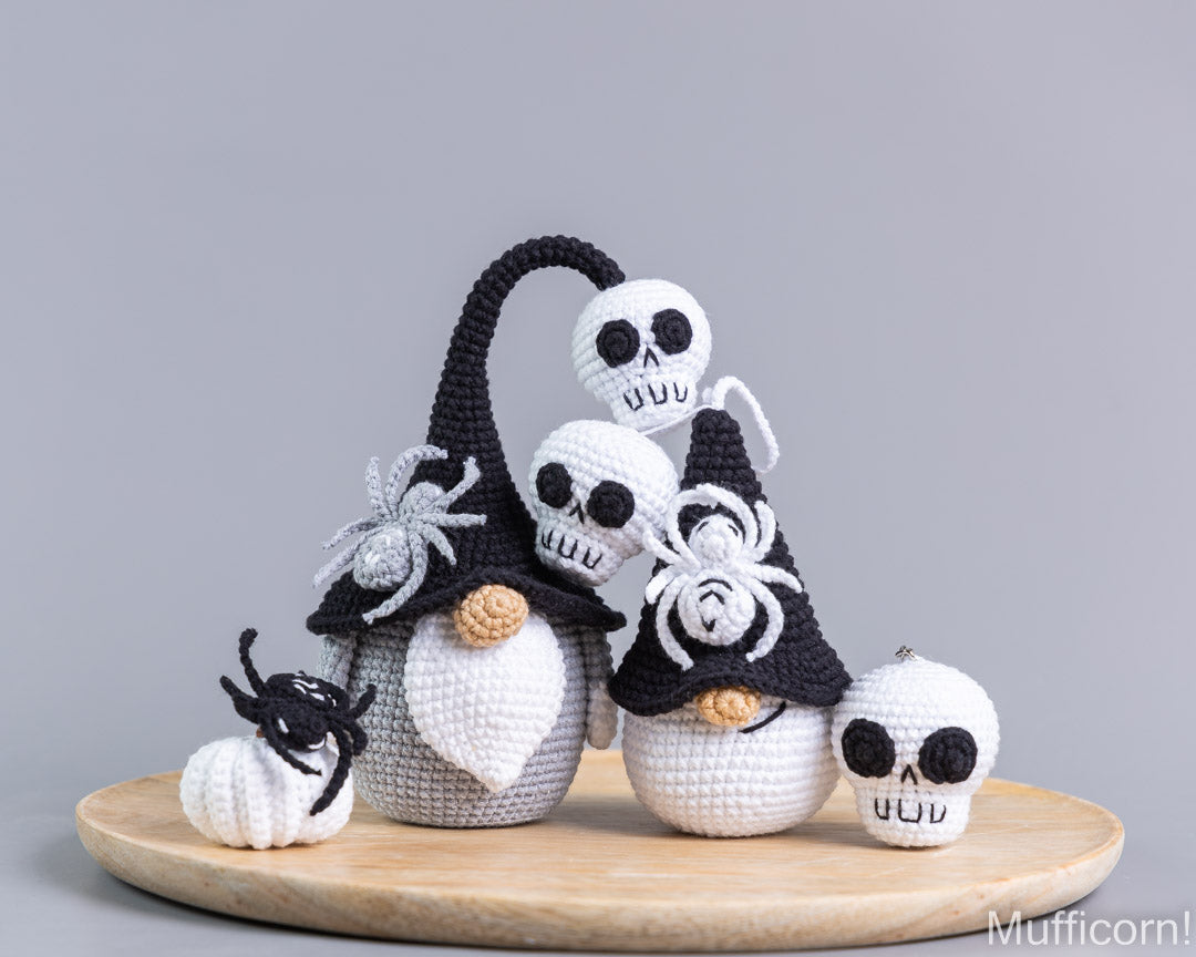A set of handmade crocheted Halloween gnomes and spider decorations displayed on a round wooden tray against a neutral gray background. The set includes three gnomes with black pointed hats, white beards, and skull faces, along with two black spiders and a small white skull.