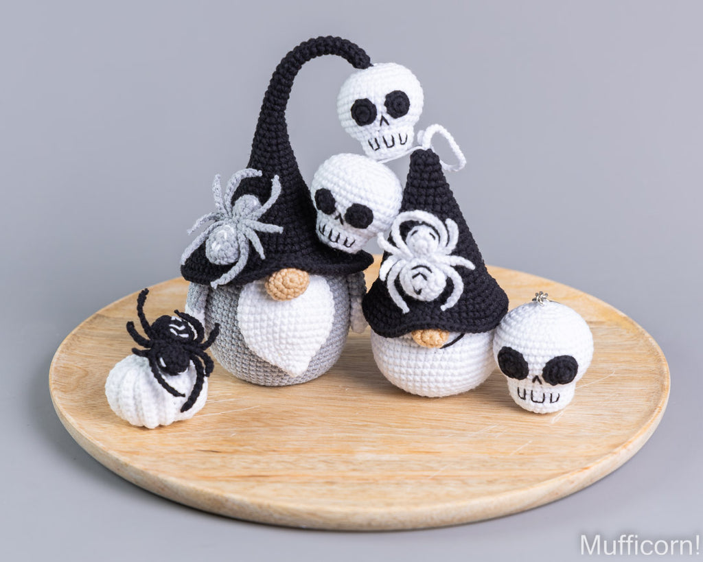 A set of crocheted Halloween gnomes and spider decorations displayed on a round wooden tray against a neutral gray background. The gnomes are white with black pointed hats, black spider details, and small beige noses.