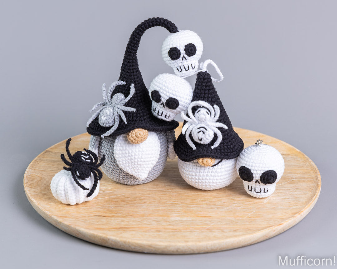 A set of crocheted Halloween gnomes and spider decorations displayed on a round wooden tray against a neutral gray background. The gnomes are white with black pointed hats, black spider details, and small beige noses.