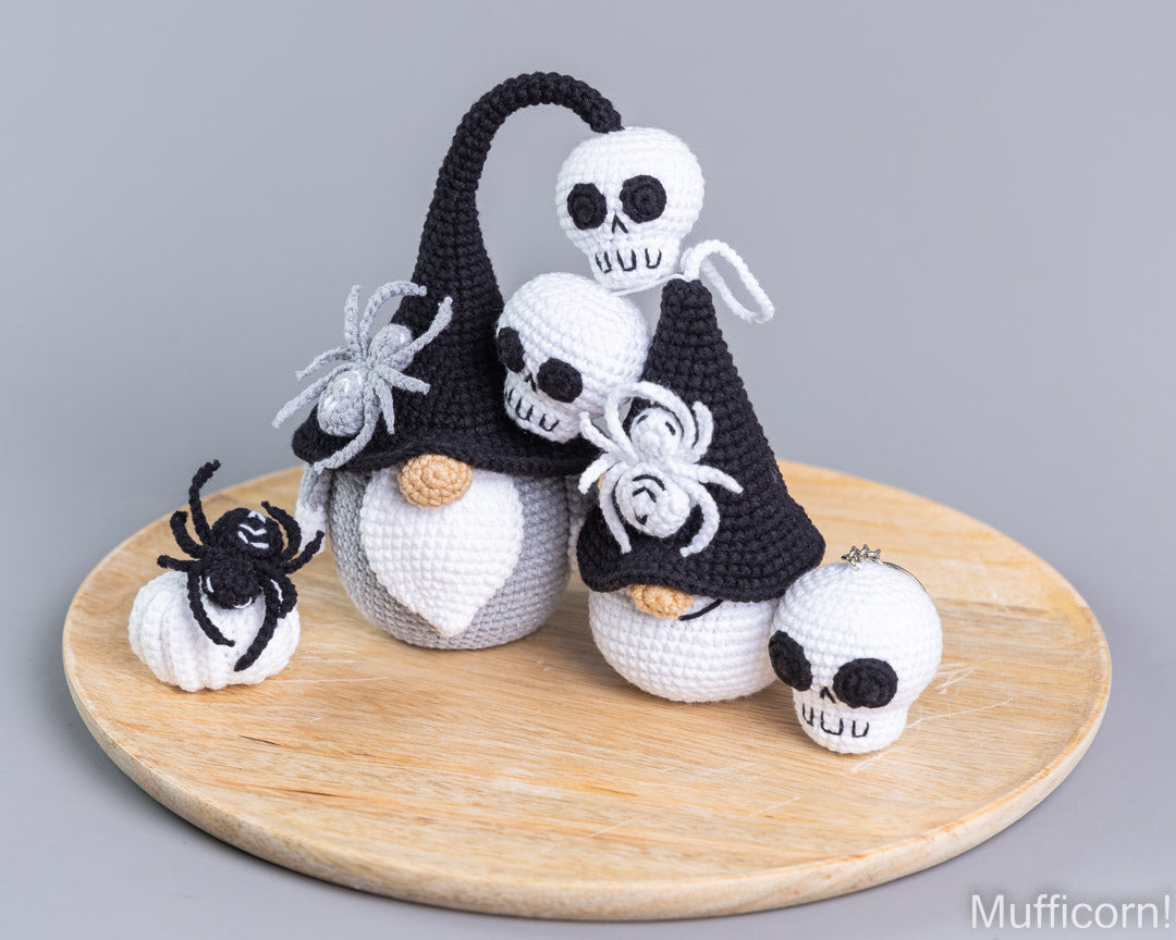 A set of crocheted Halloween gnomes styled with skulls, black pointed hats, white beards, and spiders, arranged on a round wooden board against a solid gray background.