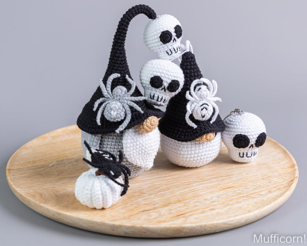 Four crocheted Halloween gnomes with skull faces and black pointed hats decorated with white spiders, arranged on a round wooden tray against a light gray background.