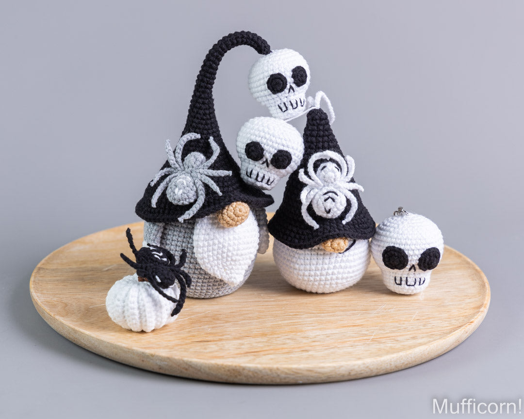 A set of crocheted Halloween decorations including three skull-shaped muffs in black and white pointed hats with embroidered spiders, a small pumpkin, and a skull ornament, displayed on a round wooden tray against a gray background.