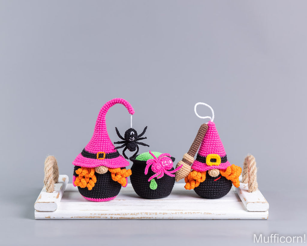 Three crocheted Halloween gnomes on a white rustic tray against a gray background. The gnomes have pink and black pointed hats, orange beards, and are decorated with a black spider, a pink pumpkin, and a brown broom.
