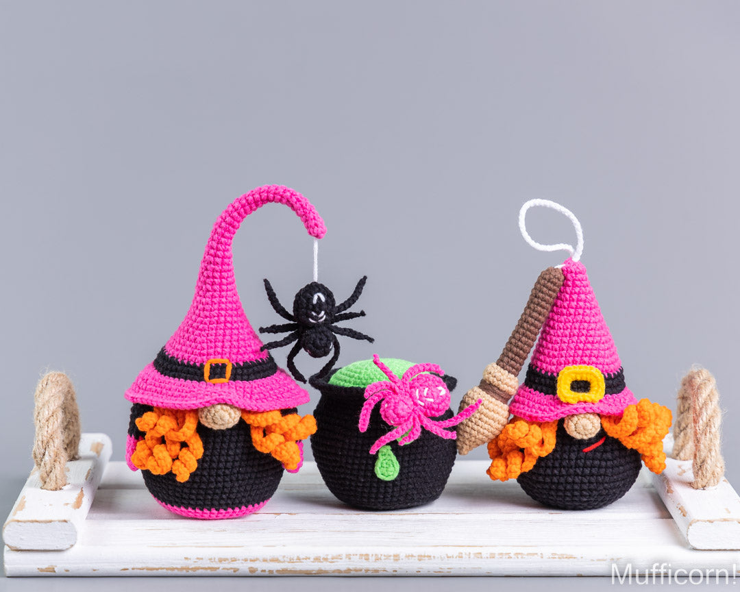 Three crocheted witch-themed decorative figures or ornaments displayed on a white wooden surface against a light gray background. The figures include black pots or hats with pink witch hats, orange fuzzy hair, and small spider decorations.