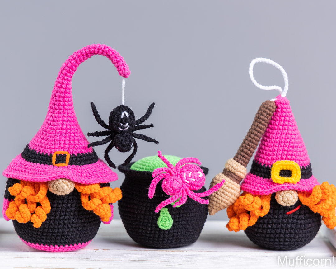 Three crocheted Halloween-themed yarn ornaments on a white surface against a gray background: a witch gnome with a curved pink hat and orange beard, a black cauldron with a pink spider decoration, and another witch gnome holding a broom. A white hanging loop is attached to the right ornament.
