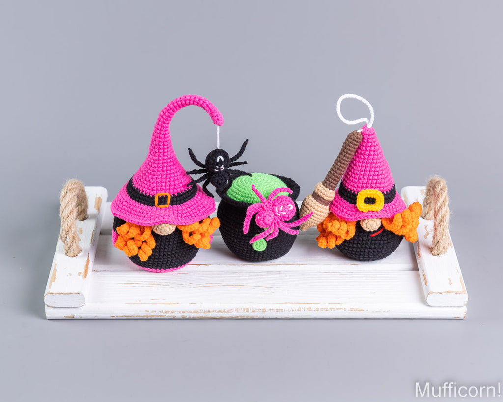 Three crocheted witch-themed ornaments displayed on a white wooden tray against a solid gray background. The ornaments feature pink pointed hats, black bodies, orange pumpkin accents, and decorative details including a black spider, a green cauldron, and a broom.