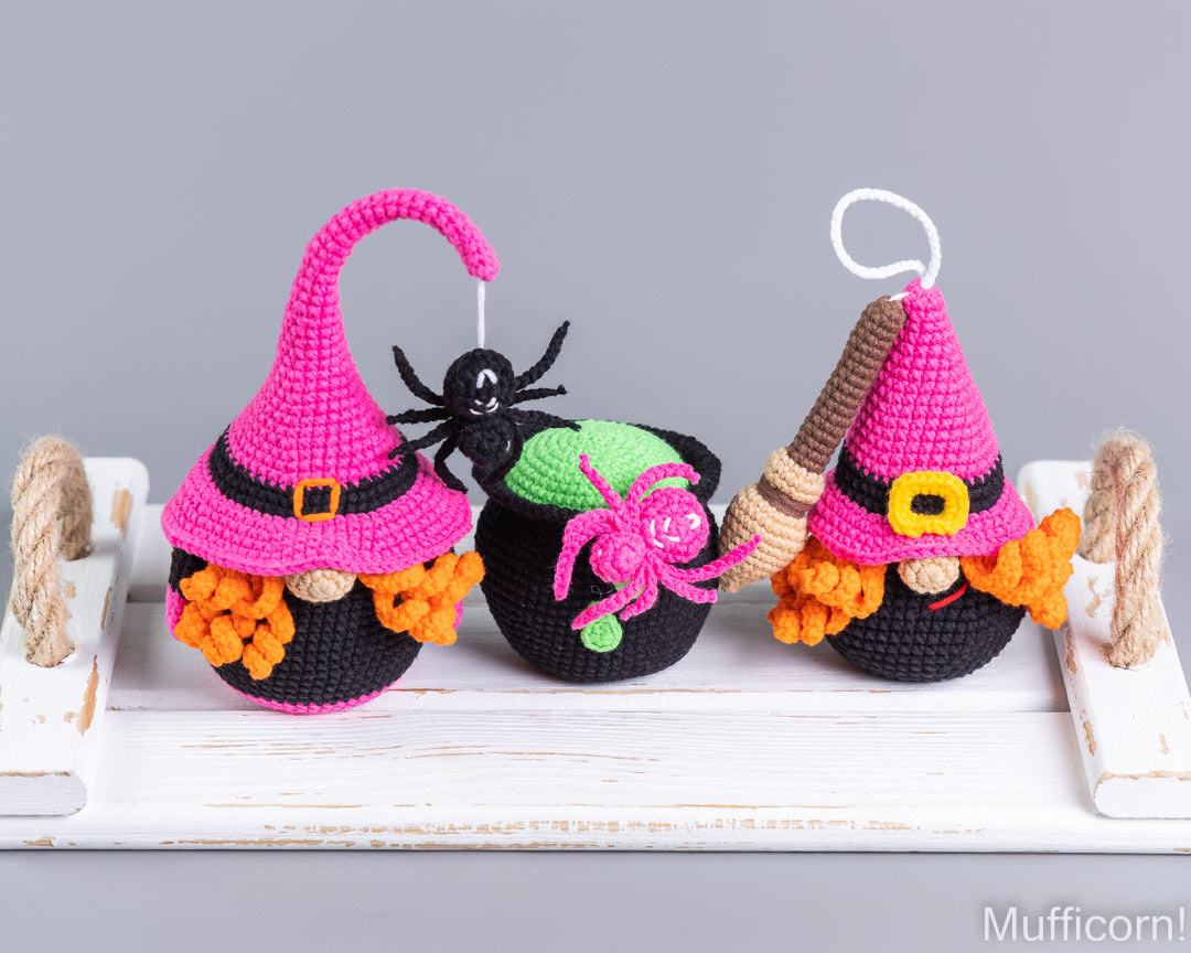 Three crocheted Halloween ornaments shaped like witch hats and cauldrons displayed on a white wooden tray against a gray background. The ornaments are black, pink, orange, green, brown, and yellow, with decorative black spiders and a broomstick.