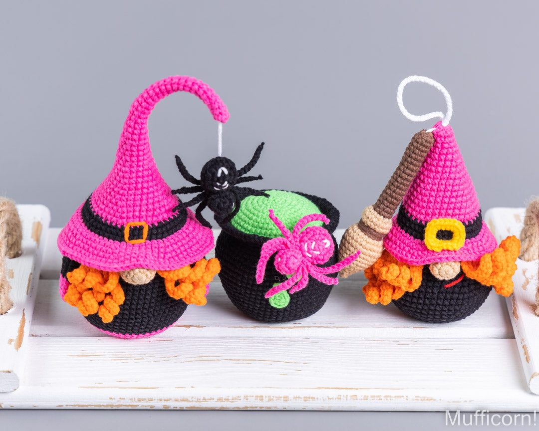 Three crocheted Halloween gnomes with pointed hats, orange yarn hair, and black yarn pants are arranged on a white wooden surface. One gnome has a pink hat and beard, one has a green cauldron on top, and one has a brown broom.