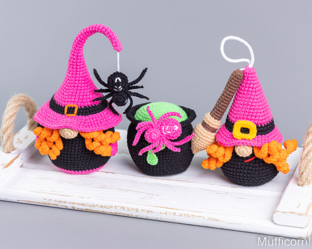 Three crocheted Halloween gnome ornaments displayed on a white wooden tray against a gray background. The left and right gnomes wear bright pink witch hats with black bands, orange yarn hair, and black bodies, while the center gnome has a black body, a green hat, and a pink spider decoration.