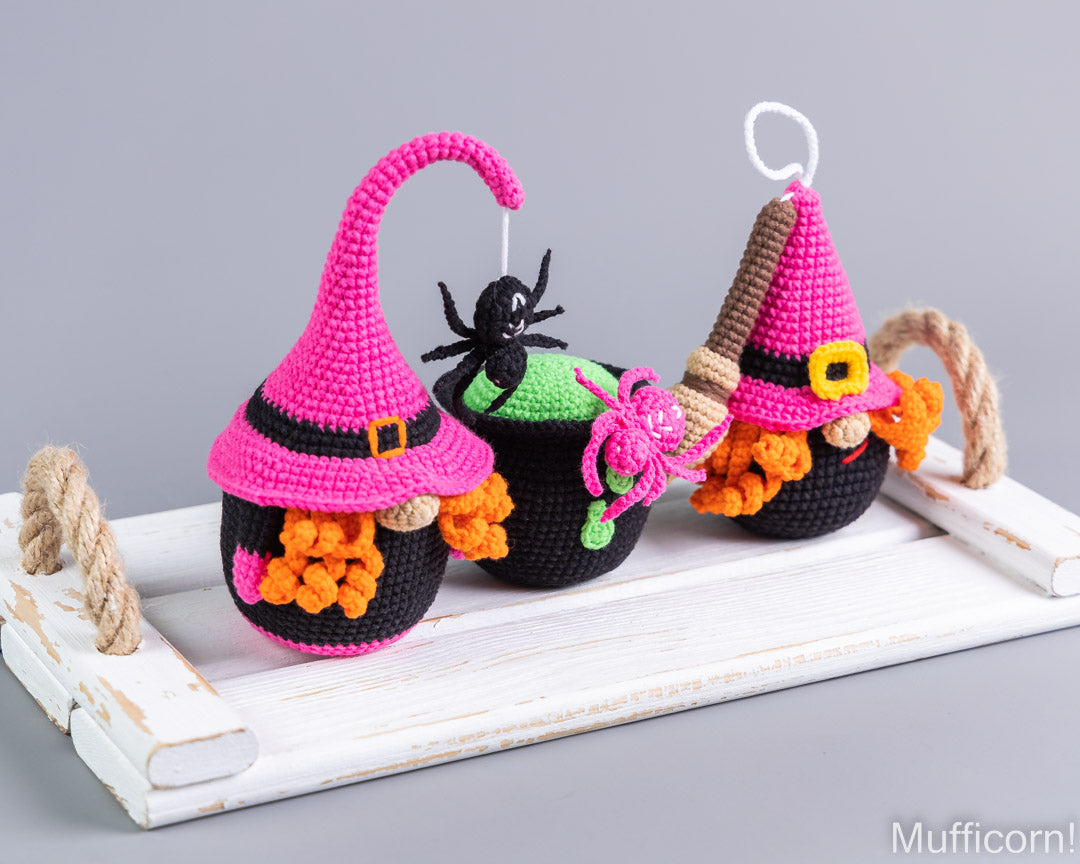 Three crocheted Halloween ornaments shaped like witch hats over pots, displayed on a white wooden tray against a gray background.