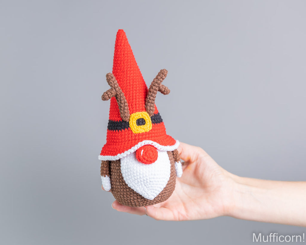 A hand holding a crocheted Christmas gnome reindeer with a red pointed hat, brown antlers, a white beard, and a red nose, set against a plain gray background.