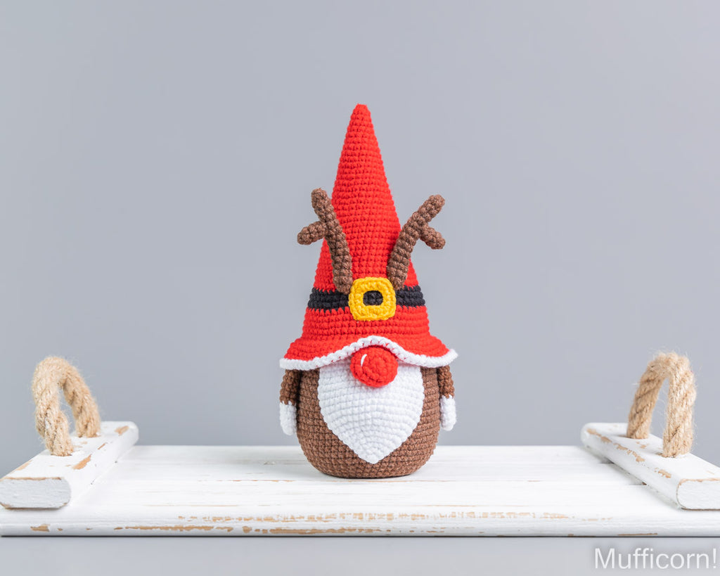 Crocheted Christmas gnome doll with a red pointed hat, brown antlers, white beard, and red nose, displayed on a white wooden surface against a light gray background.