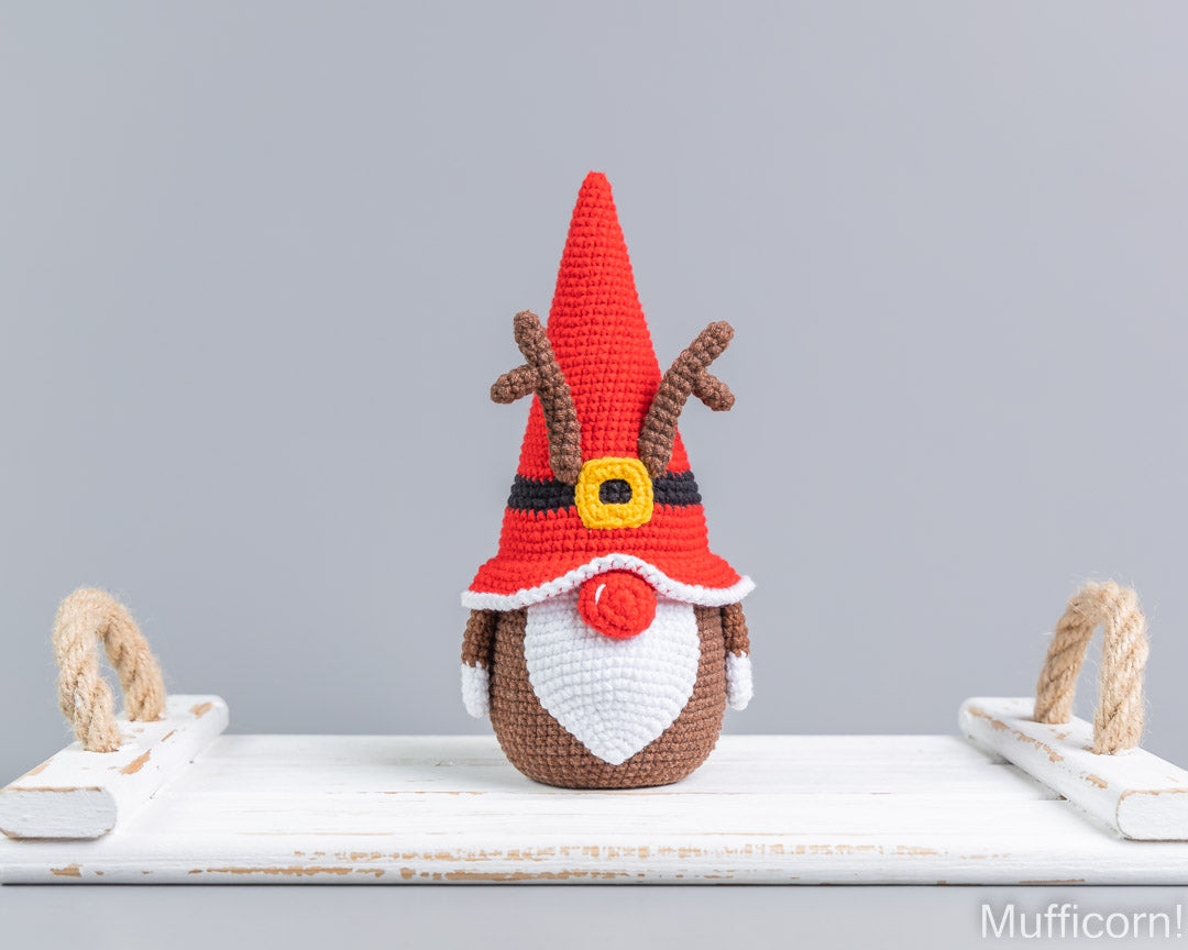 Crocheted Christmas gnome doll with a red pointed hat, brown antlers, white beard, and red nose, displayed on a white wooden surface against a light gray background.