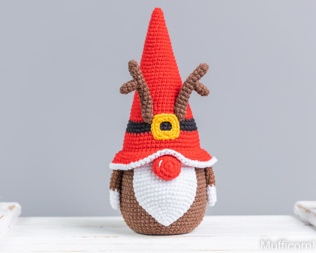 Crocheted Christmas gnome reindeer doll with a red pointed hat, brown antlers, white beard, and red nose, standing on a white surface against a light gray background.