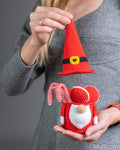 A person holds a crocheted Christmas gnome with a red and white body, white beard, and red-and-white striped candy cane, while lifting a separate red pointed hat above it. The person wears a gray knit top against a neutral gray background.