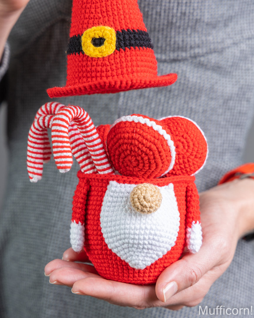 A crocheted Santa-style gnome figure in red, white, and black, holding a red-and-white striped candy cane, with a matching red hat floating above it. The figure is being held in hands against a gray knit background.