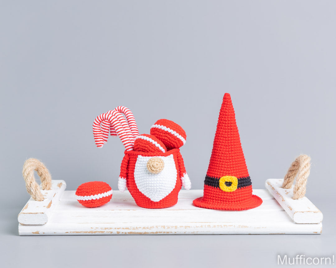 Crocheted Christmas gnome decorations displayed on a white wooden tray with rope handles against a light gray background. The set includes a Santa-style gnome with a striped red and white hat and beard, a tall red pointed hat with a black and yellow band, and a small red ball.