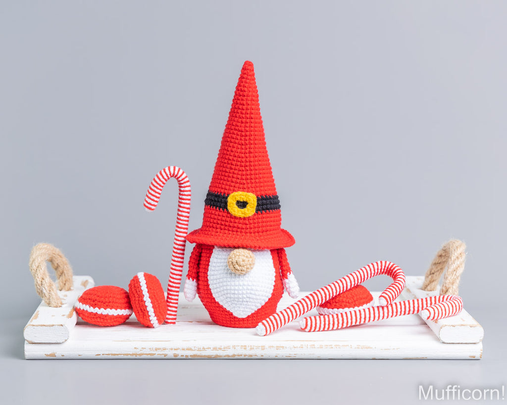 Crocheted Christmas gnome decoration on a white wooden tray, featuring a red pointed hat with a black band and gold buckle detail, a white beard, red-and-white striped candy canes, and red-and-white striped pom-poms, set against a solid gray background.