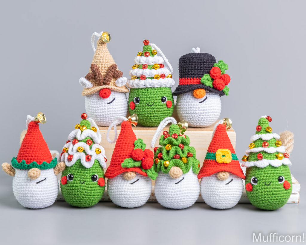 A set of nine crocheted Christmas-themed plush ornaments arranged on and around a small wooden box against a light gray studio background. The ornaments include snowman and tree designs in white, green, red, black, brown, and yellow, each with embroidered facial features and small hanging bells.