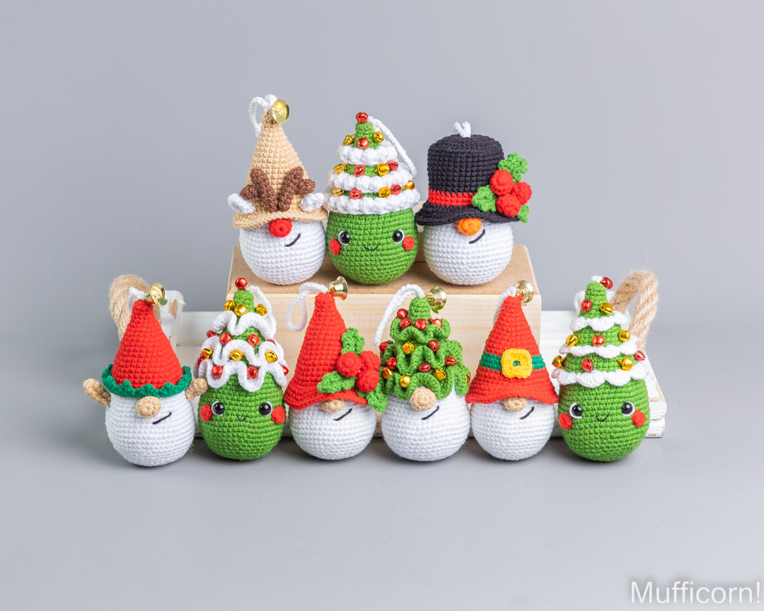 A set of crocheted Christmas-themed ornaments displayed on a light gray background, including snowman and tree characters in festive hats and outfits, arranged on a wooden block with a white box behind them.