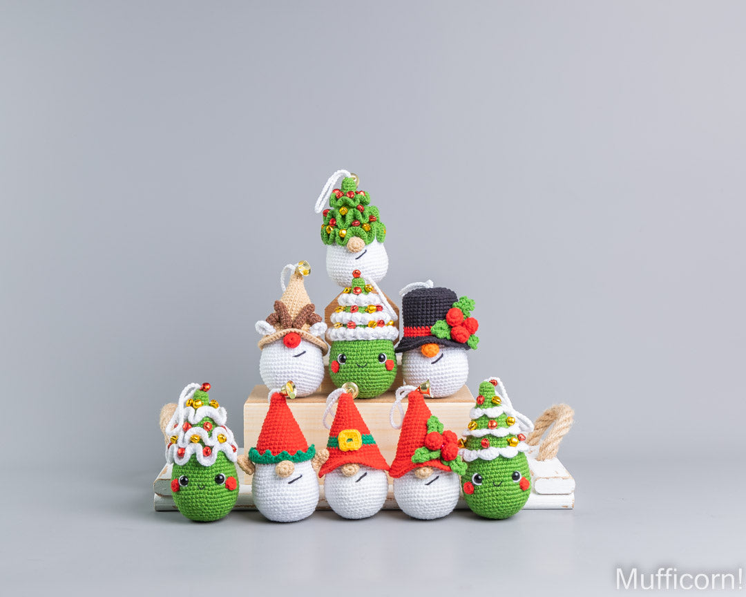 A collection of crocheted Christmas-themed amigurumi dolls arranged on a small wooden platform against a plain light gray studio background. The dolls include snowman and tree characters in red, green, white, black, and brown clothing and hats.