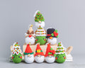 A collection of handmade crocheted Christmas ornaments shaped like snowmen wearing festive hats, arranged on a light wooden block against a solid gray background.