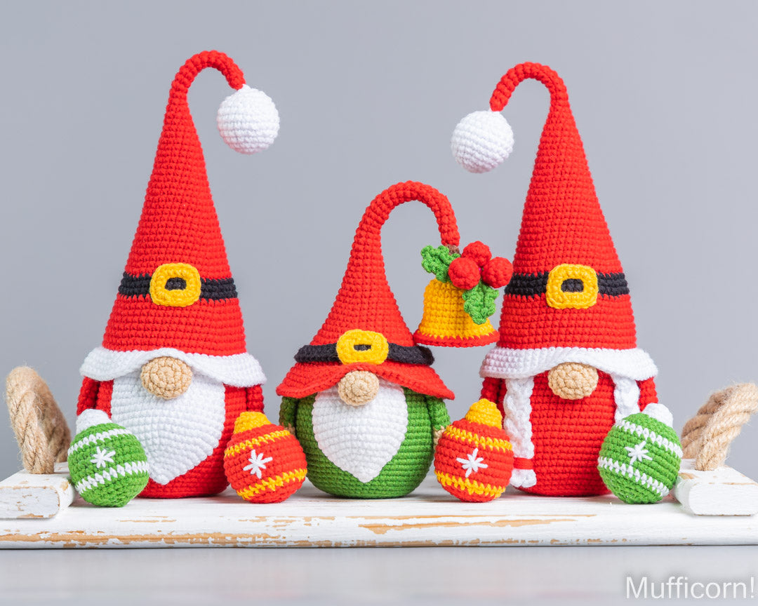 Three crocheted Christmas gnomes in red hats with white beards, arranged on a white rustic surface with colorful ornaments and a small bell, against a light gray background.