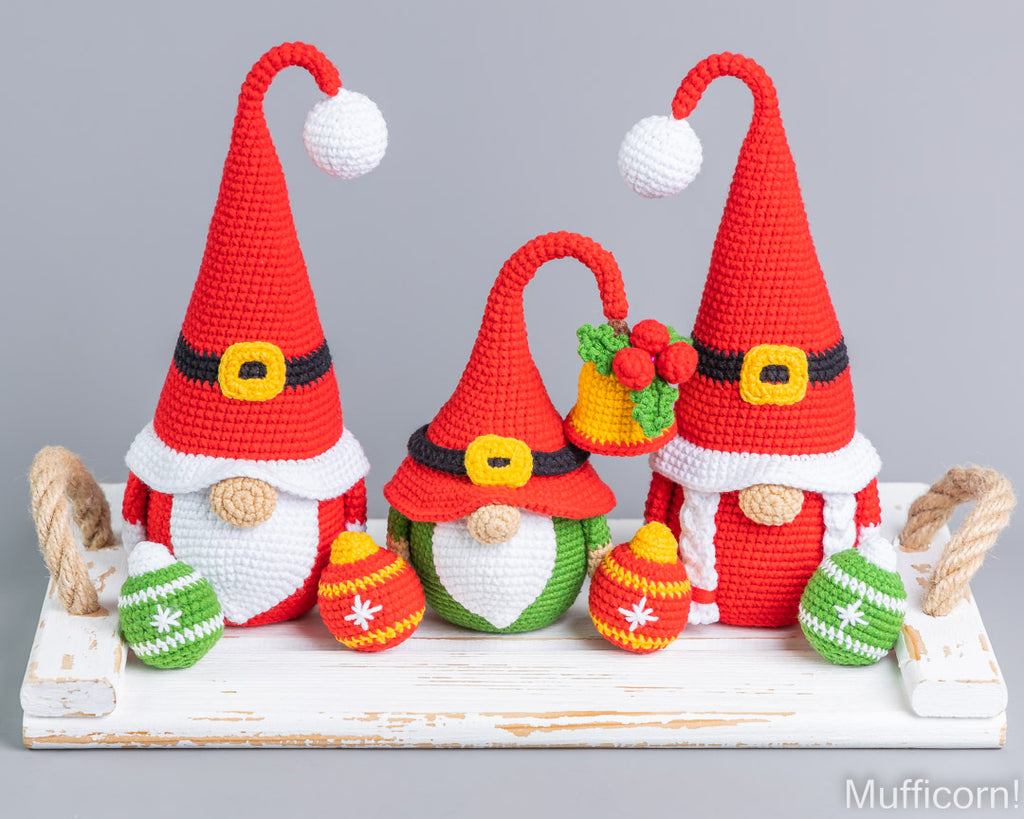 Three crocheted Christmas gnome figures arranged on a white rustic tray against a light gray background. The gnomes wear red Santa hats with white pom-poms, white beards, and black belts, with one holding a yellow bell and one decorated with green and red ornaments.