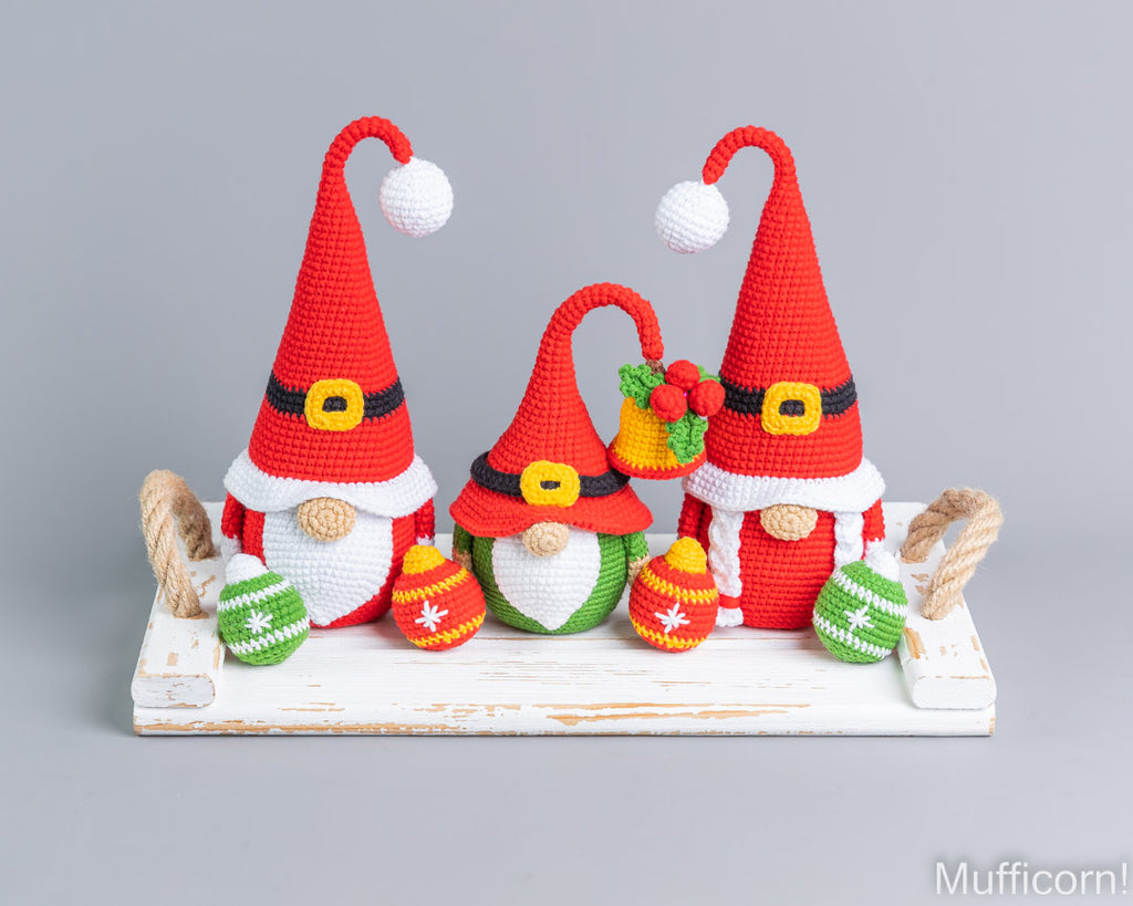 Three crocheted Christmas gnome decorations displayed on a white rustic wooden tray against a solid gray studio background. The gnomes are red and white with black and yellow accents, wearing tall pointed hats with white pom-poms and holding colorful ornaments.