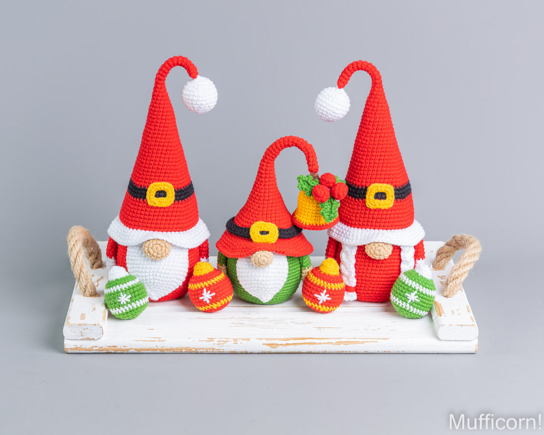 Three crocheted Christmas gnome decorations displayed on a white rustic wooden tray against a solid gray studio background. The gnomes are red and white with black and yellow accents, wearing tall pointed hats with white pom-poms and holding colorful ornaments.
