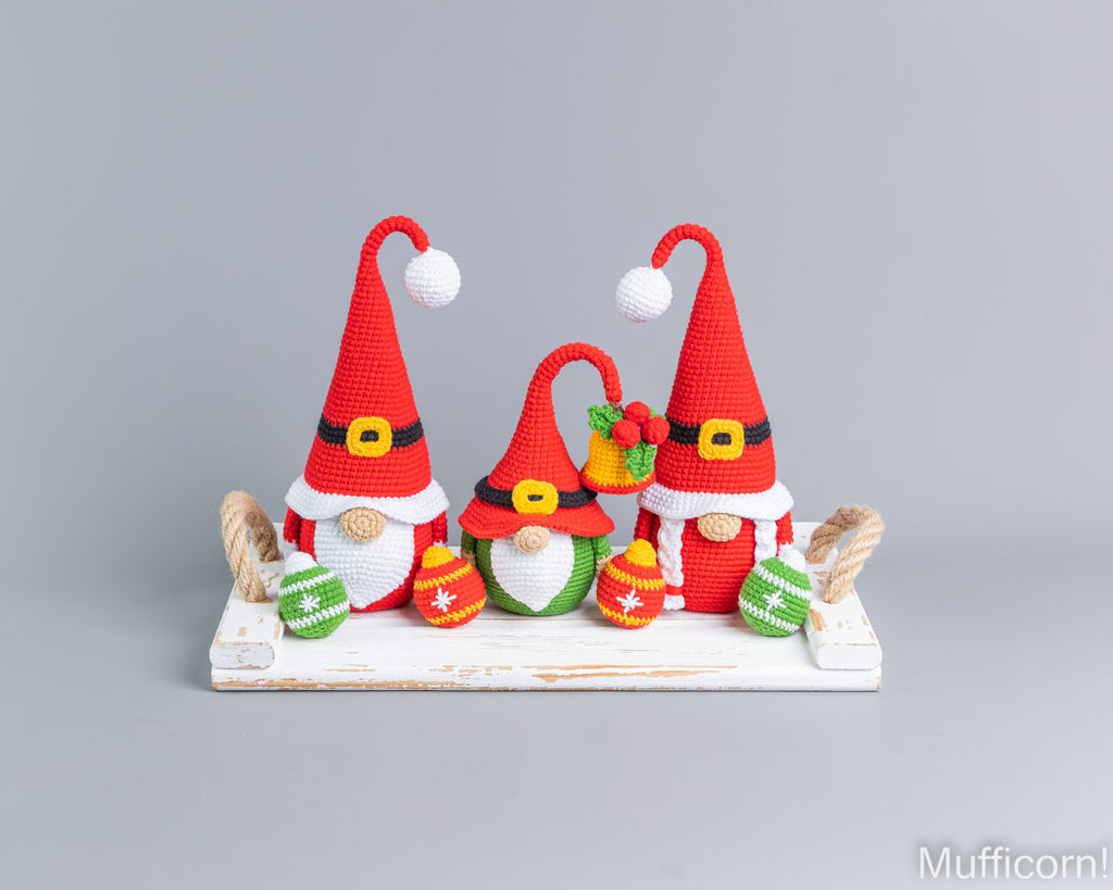 Three crocheted Christmas gnomes arranged on a white wooden base against a light gray background. The gnomes wear red hats with white pom-poms, white beards, and black-and-yellow belts, and hold colorful ornaments.