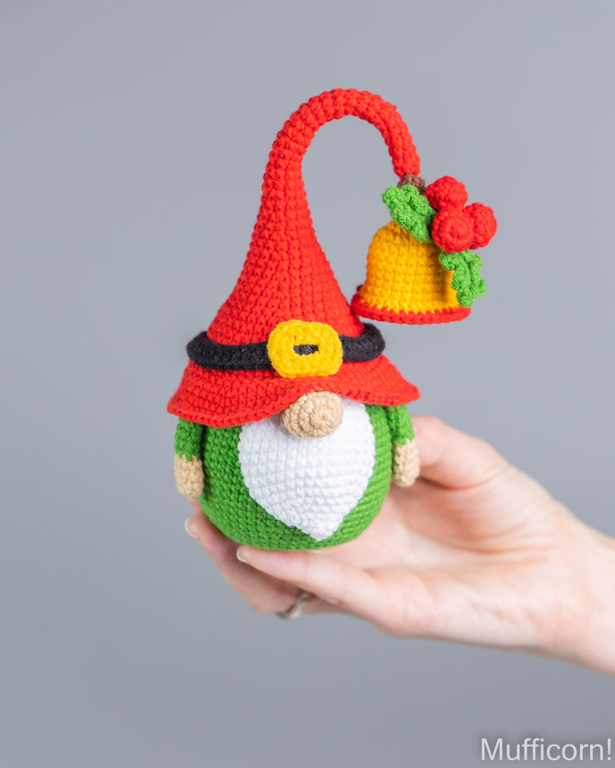 A hand holds a crocheted Christmas gnome ornament against a plain gray background. The gnome has a red hat with a black band, a yellow buckle detail, a green body, a white beard, and a yellow bell with red flowers hanging from the hat.