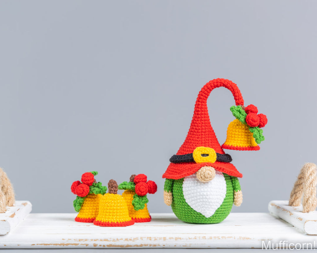 Crocheted Christmas Santa gnome decoration with a red hat, yellow bell, white beard, green body, and small red flower accents, displayed on a white surface against a light gray background.
