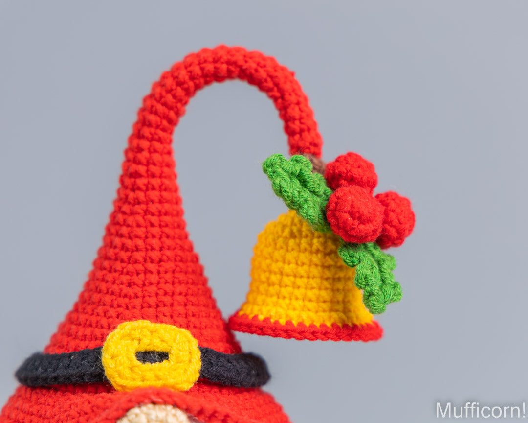 A close-up of a crocheted gnome hat in bright red, featuring a yellow bell with green leaves and red berries hanging from the brim, set against a solid gray background.