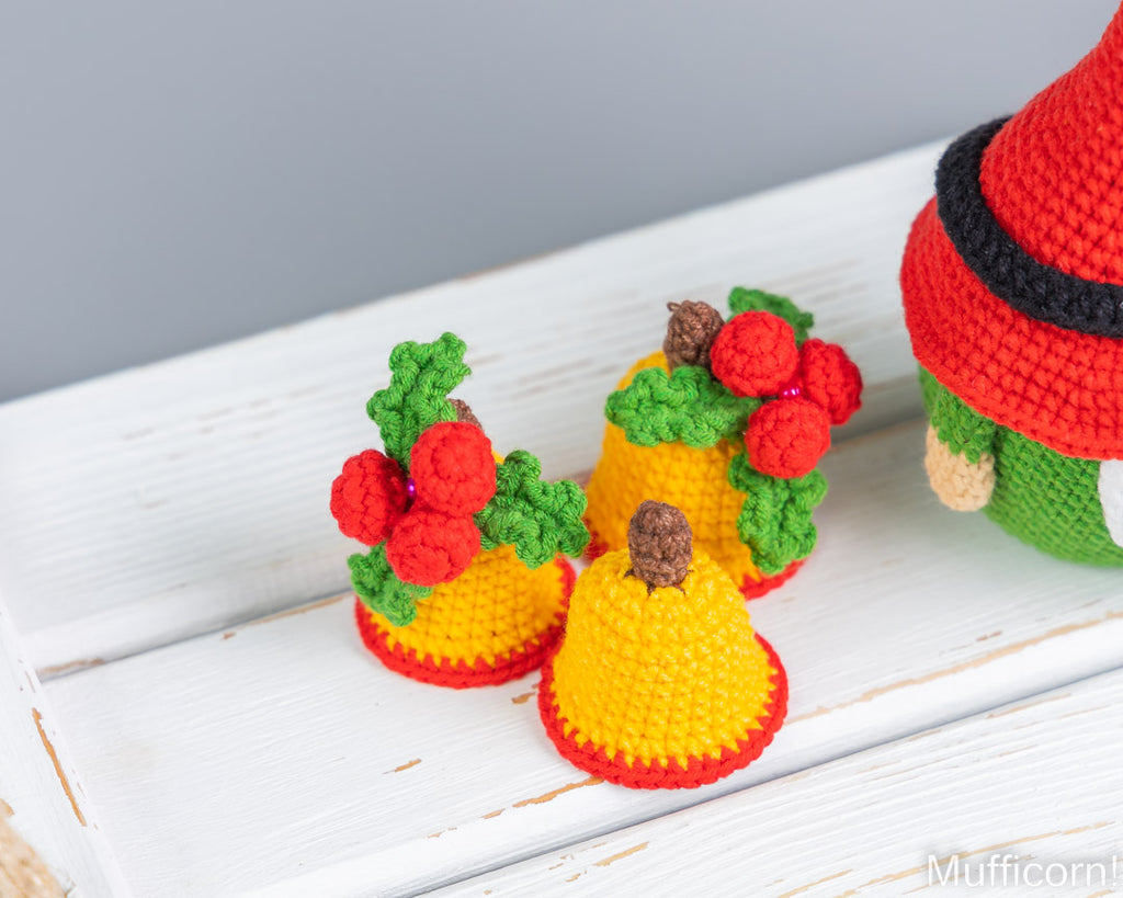 Three crocheted yellow Christmas bells with red trim and green holly with red berries, arranged on a white wooden surface next to a partially visible green and red gnome figure.