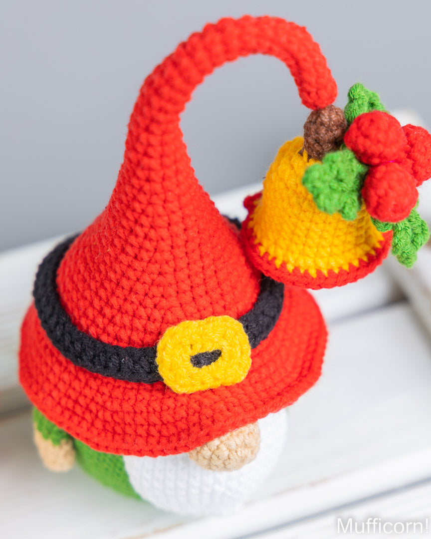A crocheted gnome figurine with a tall red hat, black belt, yellow buckle, white face, green shirt, and a yellow bell with red berries on a white surface.