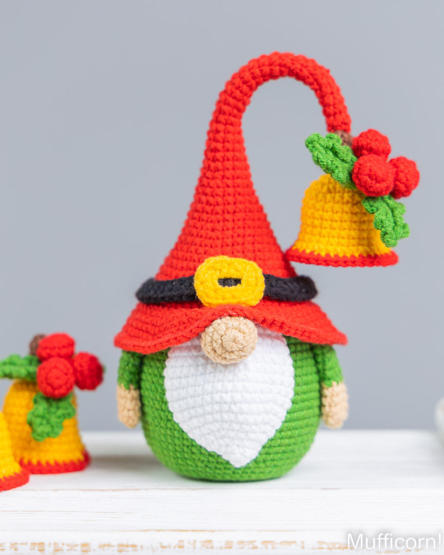 A crocheted Christmas gnome ornament with a tall red hat, white beard, green body, and yellow bell decoration, displayed on a white surface against a light gray background.