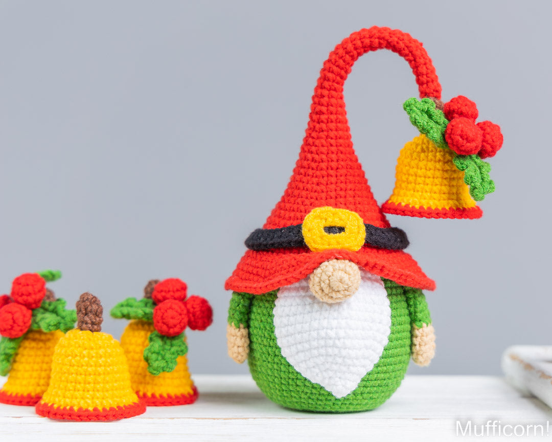 Crocheted Christmas gnome decoration with a red pointed hat, yellow bell, green body, white beard, and red nose, shown on a light surface with smaller matching ornaments in the background.