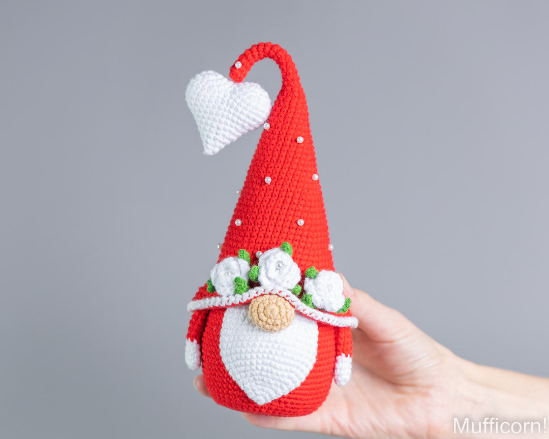A crocheted gnome doll with a red pointed hat, white beard, and white flowers, held in a person's hand against a plain gray background.