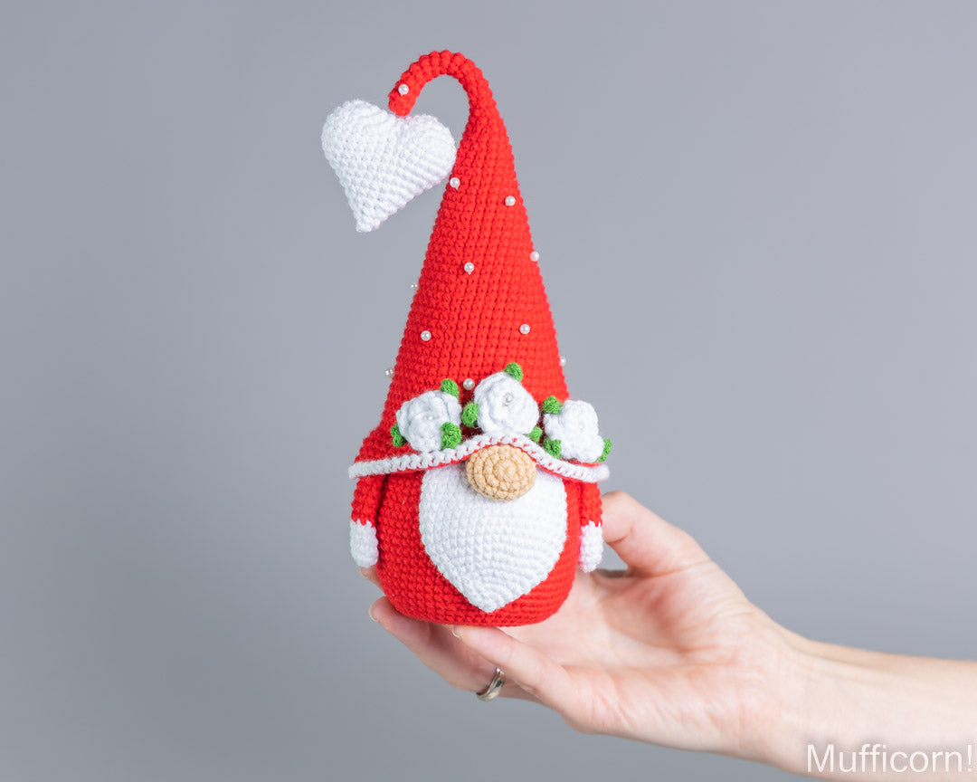 A crocheted gnome doll with a red pointed hat, white beard, and small green and white floral details, being held in a person's hand against a plain gray background.