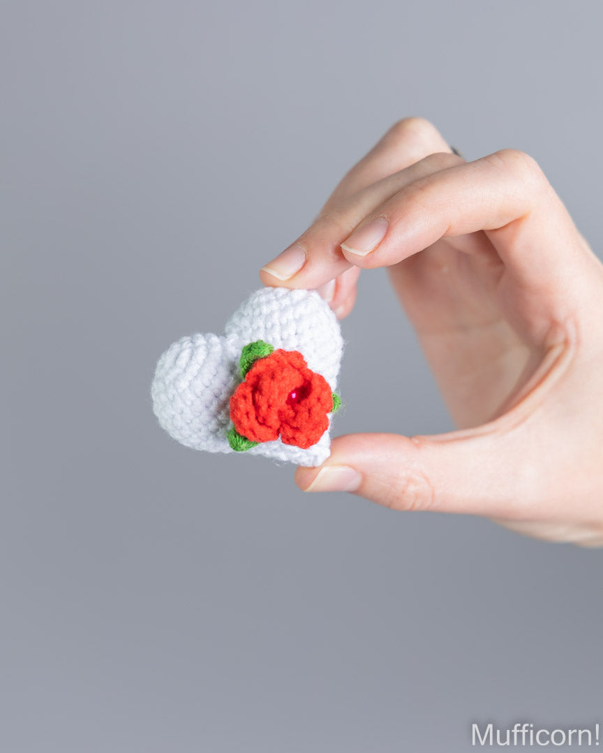 A hand holds a small crocheted white heart with a red rose and green leaves against a plain gray background.
