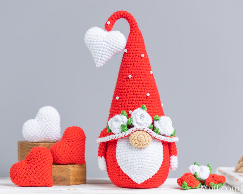 A crocheted gnome decoration with a tall red hat, white heart-shaped face, white flowers, green leaves, and small white bead details, displayed on a tabletop with red and white heart props against a light gray background.